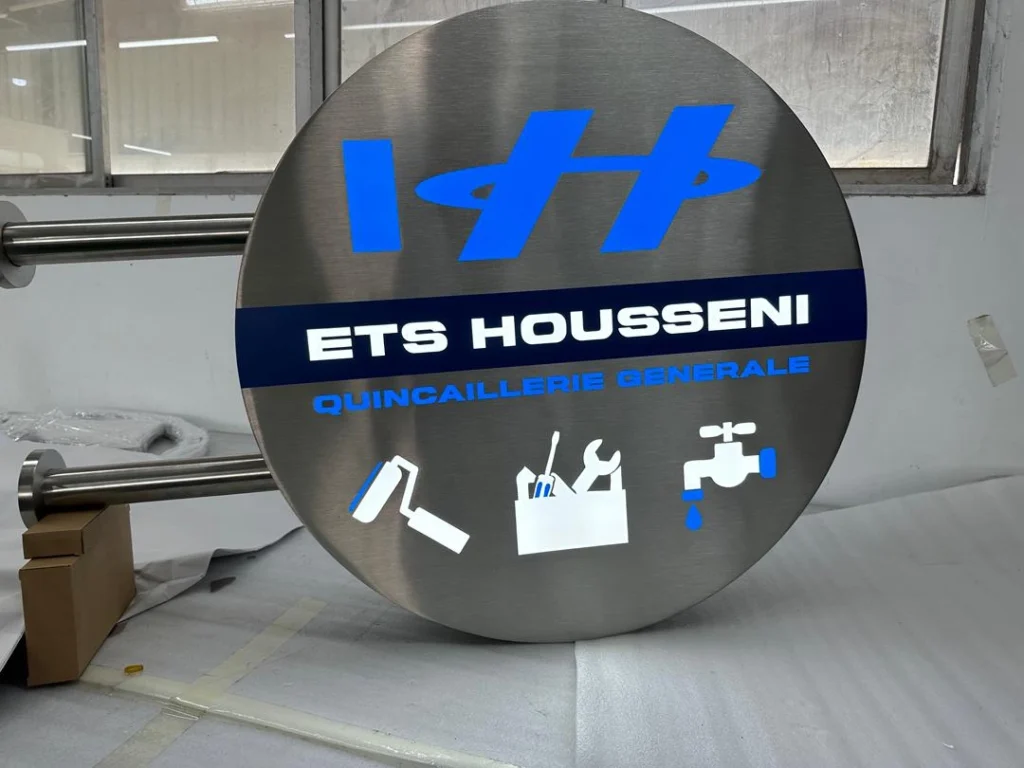 Enseigne drapeau lumineuse inox - ETS Housseni Enseigne drapeau ronde en inox brossé avec lettrage lumineux bleu et blanc pour ETS Housseni quincaillerie générale