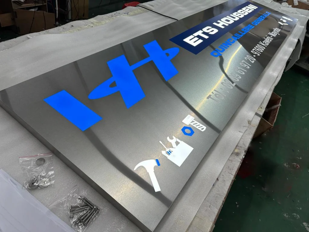 Enseigne tôle ajourée inox - ETS Housseni Enseigne en tôle ajourée inox brossé avec lettrage bleu et blanc réalisée pour ETS Housseni quincaillerie générale